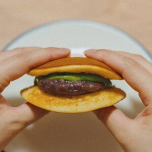 Hands holding a small hamburger with a beef patty and green toppings between two buns over a plate, shown in close-up.