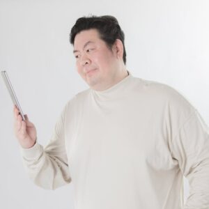 Man in a light beige turtleneck examining a tablet held in his left hand, standing against a white background.