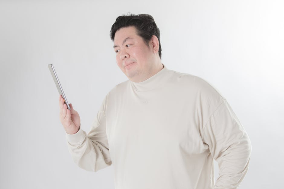 Man in a light beige turtleneck examining a tablet held in his left hand, standing against a white background.