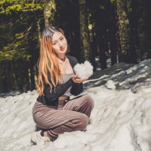 Young woman with long orange hair kneeling in a snowy forest, holding a snowball toward the camera.