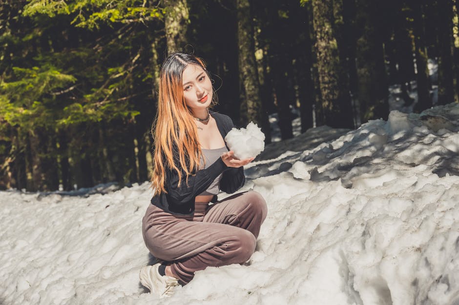 Young woman with long orange hair kneeling in a snowy forest, holding a snowball toward the camera.