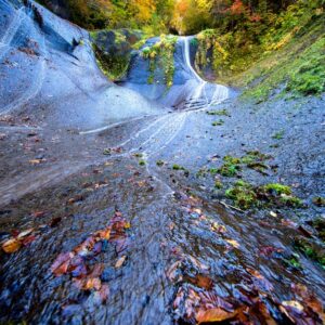 Blue-gray rock channel with water flowing down a slope, autumn leaves scattered on the wet surface and green foliage at the top of the ravine.