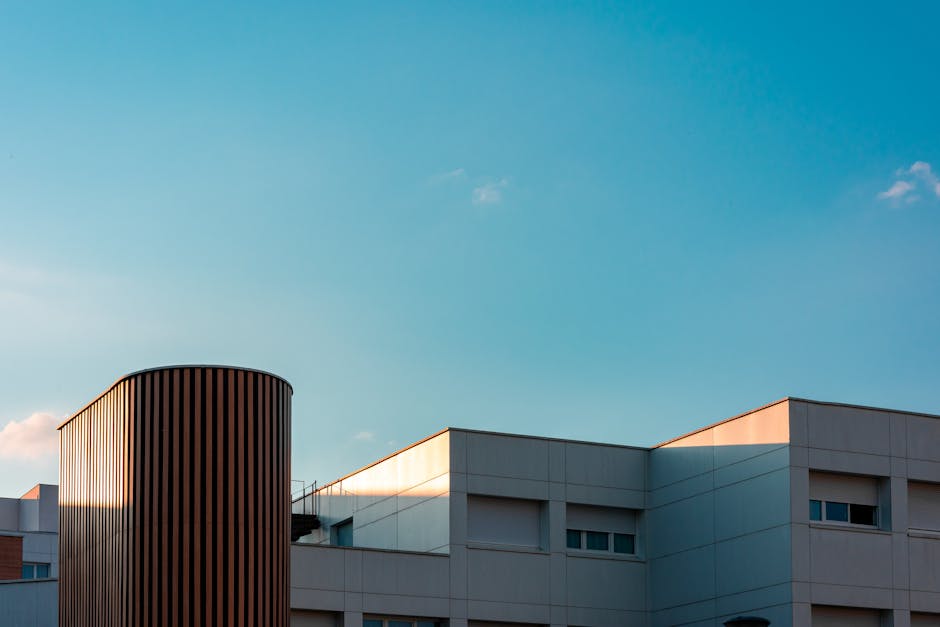 Modern white building with a tall cylindrical brown vent against a clear blue sky, early evening light casting warm highlights on the roof edges.